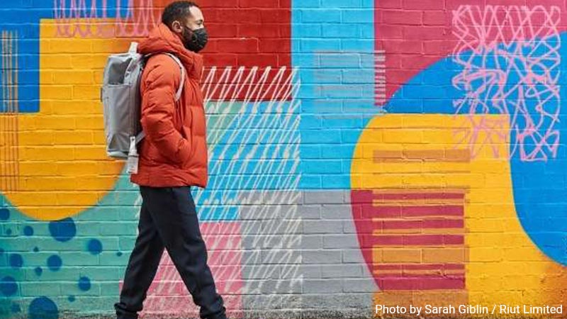 Man in a coat wearing a Riut backpack walking past a mural.